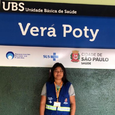 Foto de Clarice Honório.  Em pé, diante da fachada da UBS onde trabalha, Clarice está sorrindo diante da placa azul e branca com o nome Vera Poty em letras grandes. Ela tem cabelos negros lisos e usa colete azul, um crachá pendurado com fita laranja no pescoço e usa pulseiras de artesanato nos dois braços